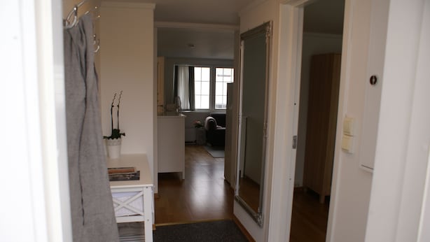 Un couloir mène à une salle de bain équipée d’un miroir, d’un porte-serviettes et d’une petite étagère où sont posés des livres et une plante.