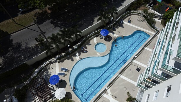Vue aérienne d’un espace piscine à la forme incurvée, entouré de chaises longues et de parasols, et situé à côté d’un bâtiment moderne.