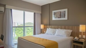 Une chambre d’hôtel avec un grand lit, deux tables de chevet avec des lampes, une œuvre d’art encadrée au mur et une vue sur la verdure par la fenêtre.