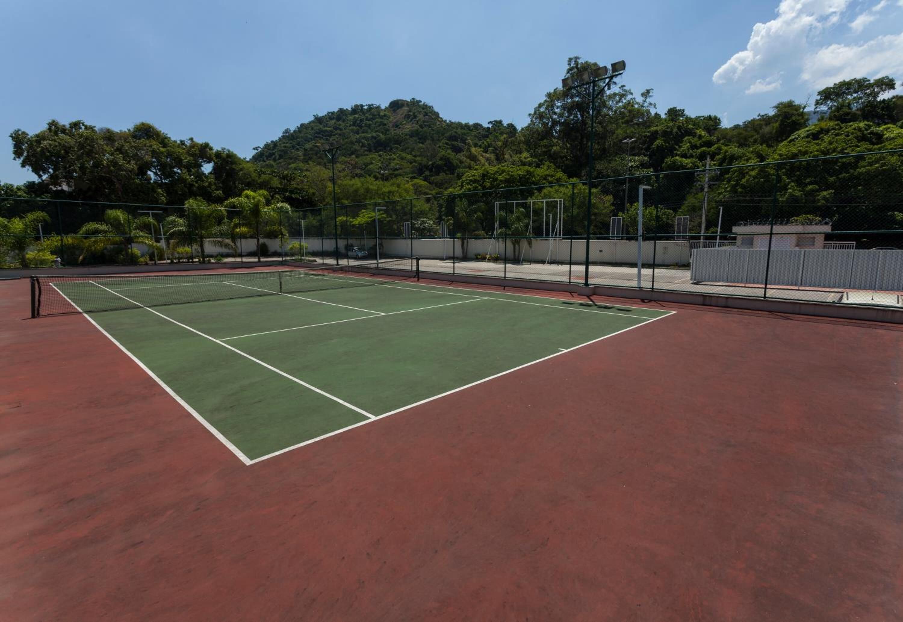 A tennis court with a green playing surface and red surrounding area, surrounded by a fence and trees.
