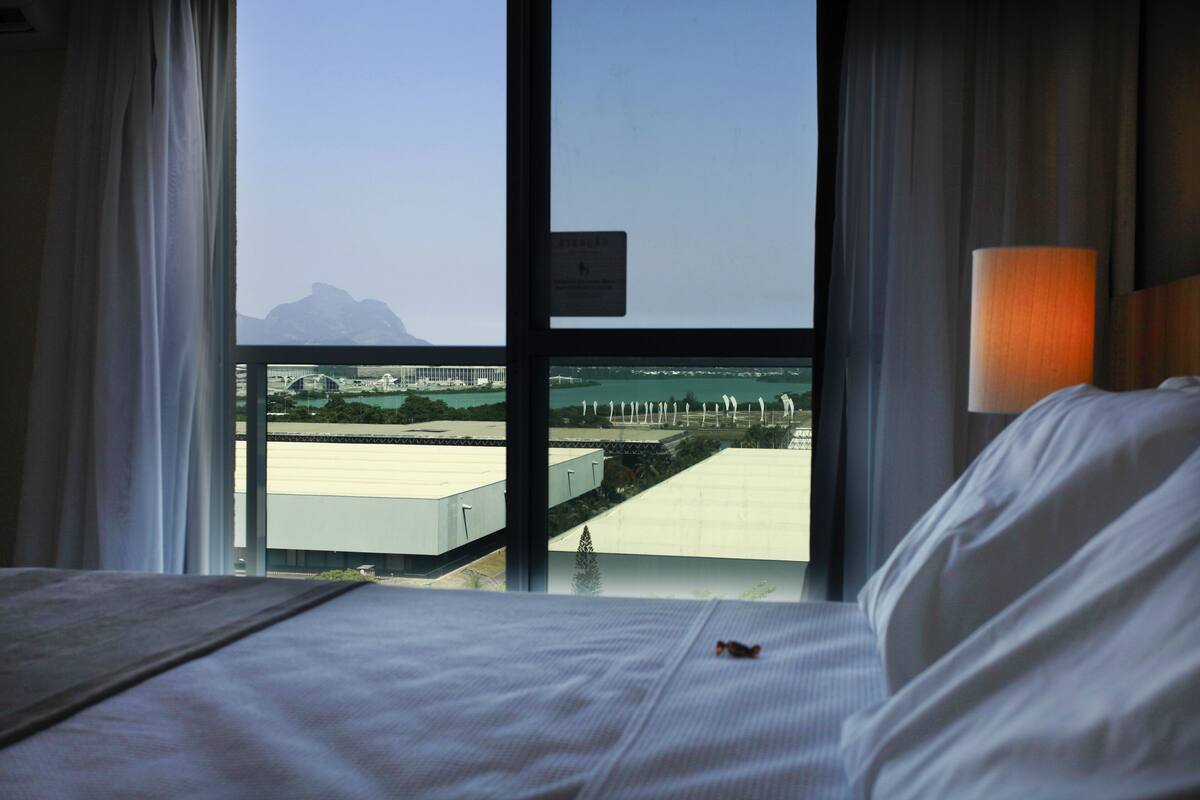 A hotel room with a bed, a window with a view of a bridge and mountains, and a lamp on the nightstand.