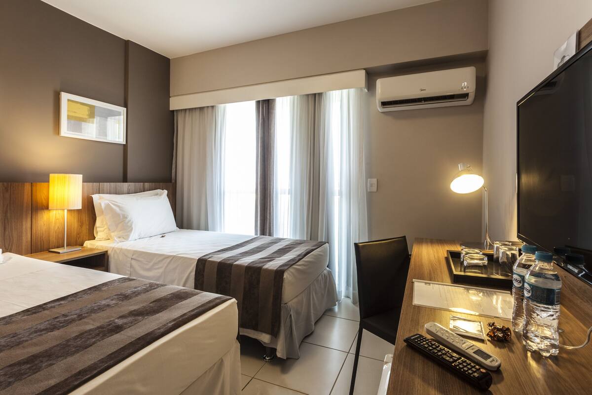 A hotel room with two beds, a desk with a lamp, a TV, and a window with curtains.