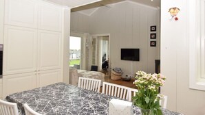 Comedor con mesa de mármol y sillas blancas, televisor montado en la pared y vistas al exterior.
