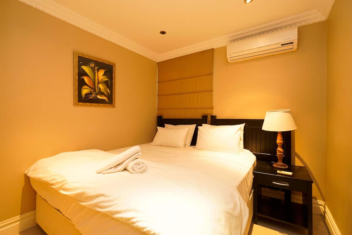 A hotel room with a bed, a nightstand with a lamp, an air conditioner, and a framed picture on the wall.