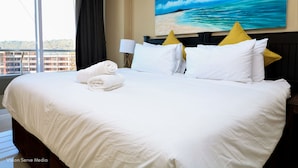Ett hotellrum med en stor säng, vita sängkläder, en tavla som föreställer en strandscen och utsikt över byggnader genom fönstret.
