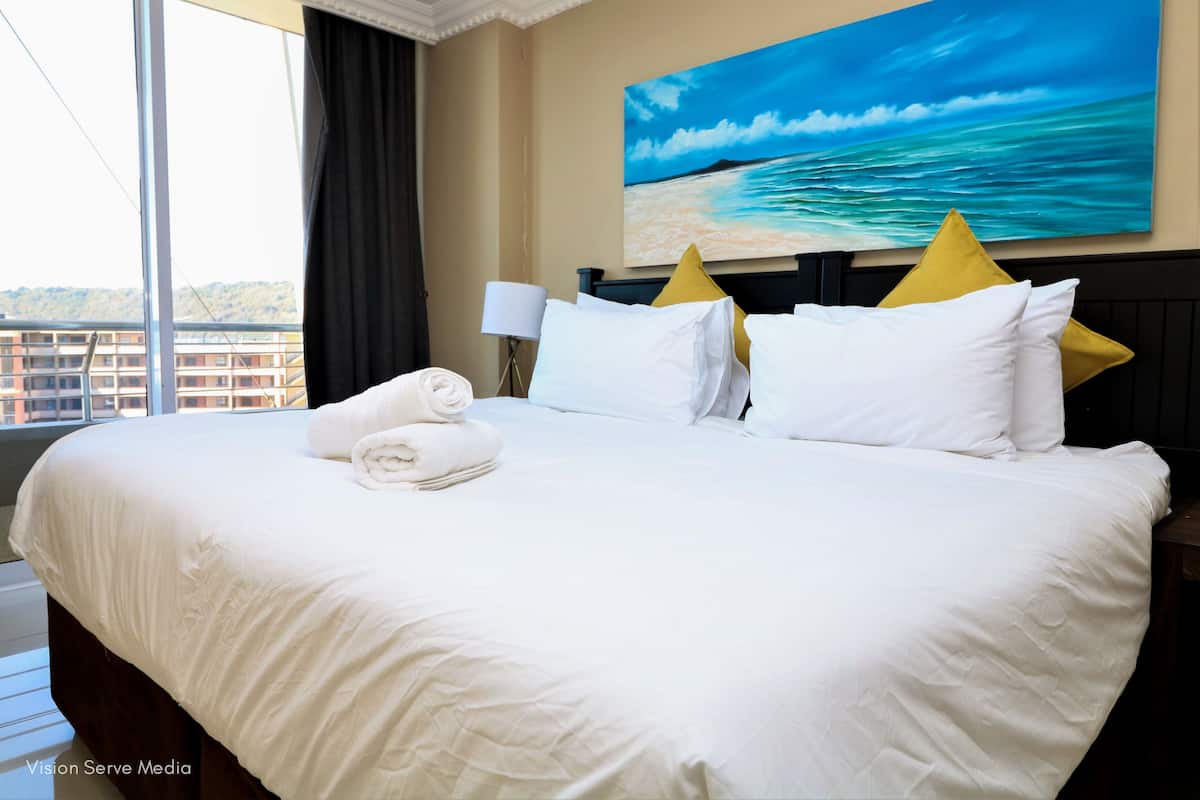 A hotel room with a large bed, white linens, a painting of a beach scene, and a view of buildings through the window.