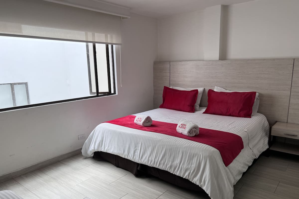 Ein modernes Hotelzimmer mit einem großen Bett, roter und weißer Bettwäsche und Blick auf die Stadt durch das Fenster.