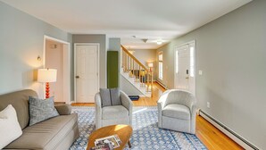 Ein Wohnzimmer mit einer Couch, zwei Sesseln, einem Couchtisch und einem gemusterten Teppich. Eine Treppe mit Geländer führt in einen anderen Raum.