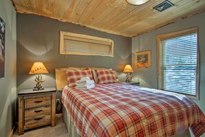 A bedroom with a plaid bedspread, two bedside lamps, a wooden headboard, a window with blinds, and a small nightstand with drawers. - Timber Home: Walk to Slopes & N Conway Shops! (North Conway)