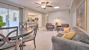 Ein modernes Wohnzimmer mit einem grauen Sofa, einem Esstisch mit Stühlen, einem Deckenventilator und einer Küche im Hintergrund.