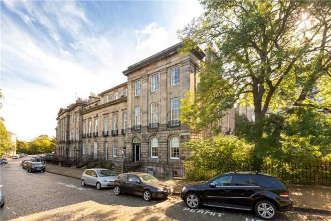 A historic building with a cobblestone street, parked cars, and trees.