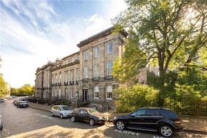 A historic building with a cobblestone street, parked cars, and trees.