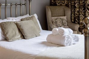 Una cama bien hecha con sábanas blancas y toallas enrolladas, un sillón estampado con un diseño de león, y una cortina estampada al fondo.