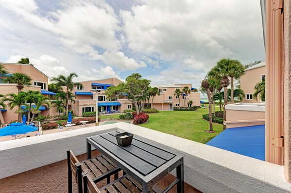 A balcony with a table and chairs, overlooking a pool and a well-maintained lawn with palm trees.