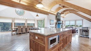 Una cocina amplia con una isla grande, armarios de madera y una salpicadura de piedra. Grandes ventanales ofrecen vistas al exterior.