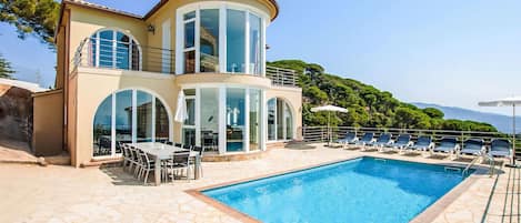 A two-story house with a swimming pool, outdoor seating, and a view of the mountains.