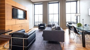 En moderne stue med et fladskærms-tv monteret på en træpanel, en grå sofa, en sort lædersofa og et spiseområde med et glasbord og stole.