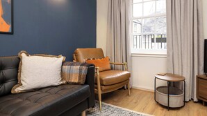 Una sala de estar con un sofá de cuero oscuro, un sillón de cuero marrón, una mesita de madera redonda, un tapiz tejido gris y una cama para mascotas gris.