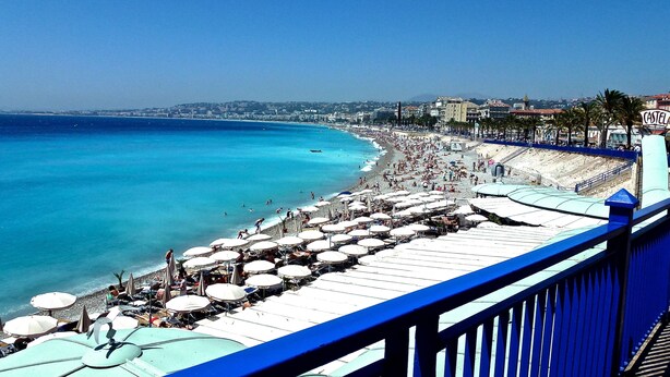 Une plage avec de nombreux parasols et des gens, une rambarde bleue et des bâtiments en arrière-plan.