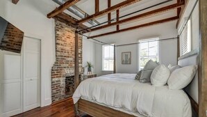 Een slaapkamer met zichtbare houten balken, een bakstenen muur, een groot bed en een raam met jaloezieën.