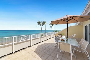 A balcony with a table, chairs, and an umbrella overlooking a beach with palm trees and the ocean. - Location, location, location - 7 Mile Beach Oceanfront 3BR at Sunset Cove! (Grand Cayman SC302)