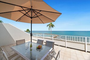 A table with a glass top, two wine glasses, and a fruit bowl, under a large beige umbrella, overlooking a beach with a palm tree and the ocean. - Location, location, location - 7 Mile Beach Oceanfront 3BR at Sunset Cove! (Grand Cayman SC302)