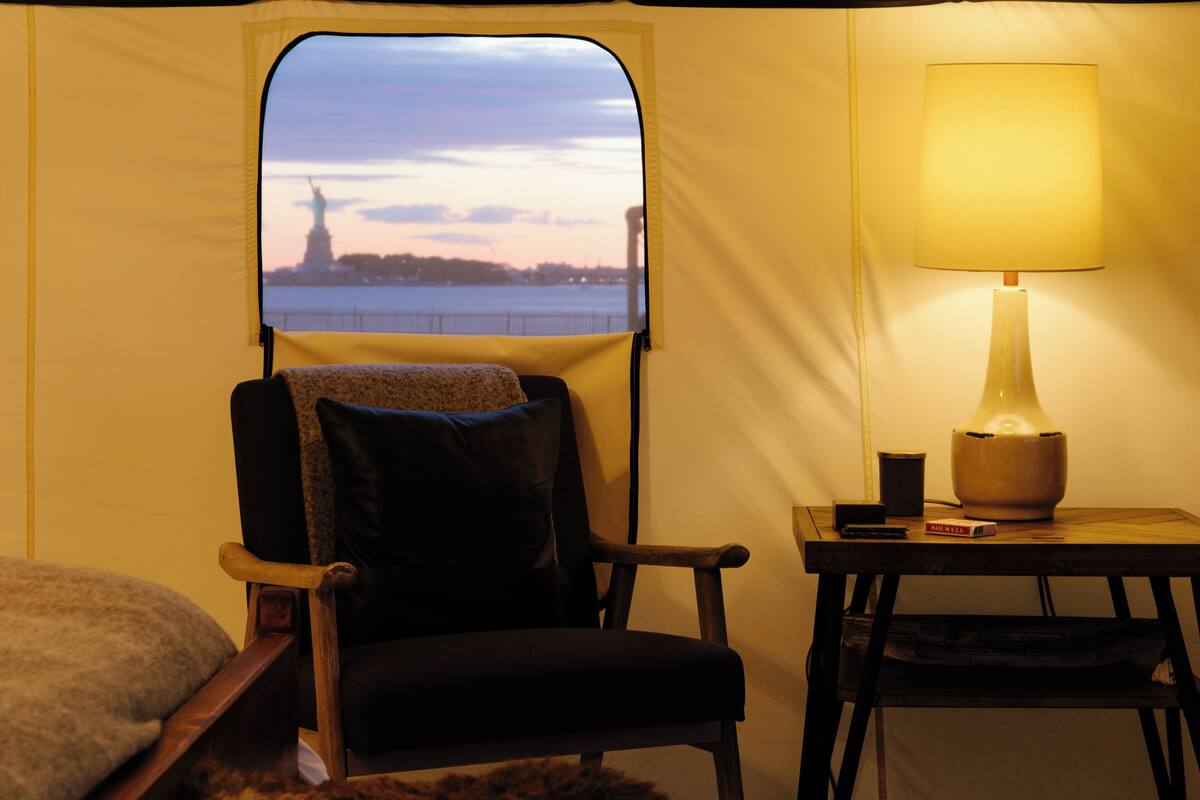 Una habitación con toldo que ofrece vista a la Estatua de la Libertad, una silla, una mesa con lámpara y un libro.