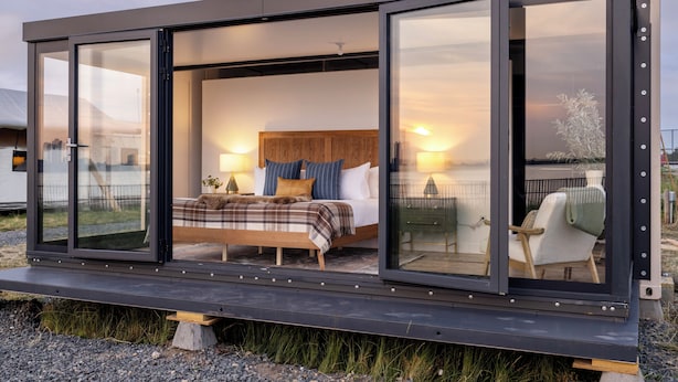 A modern, glass-walled bedroom with a wooden bed, pillows, and a nightstand. - Collective Retreats Governors Island (New York)