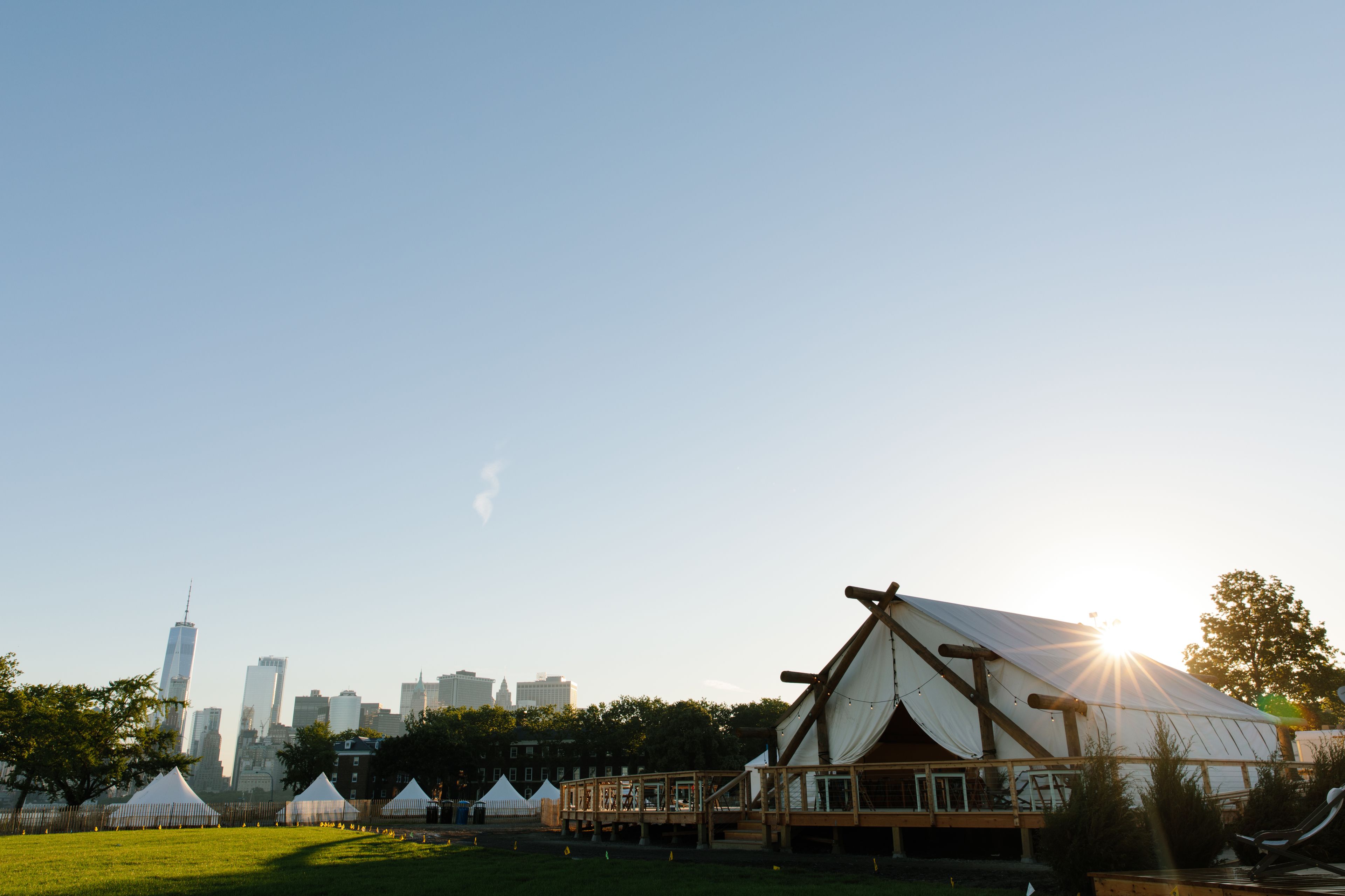 A large tent with a wooden deck, surrounded by a grassy field and trees, with a city skyline in the background.