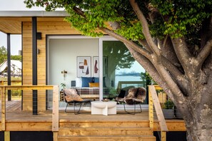 A wooden deck with outdoor seating, a glass door leading to a living area, and a large tree in the background. - Collective Retreats Governors Island (New York)