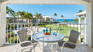 Un balcon avec une table dressée pour deux personnes, donnant sur un espace piscine agrémenté de palmiers et offrant une vue sur l’océan.