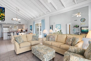 A spacious living area with a large beige sofa set, a matching ottoman, and a ceiling fan. A kitchen is visible in the background with a dining area.