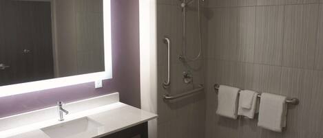 A bathroom with a sink, mirror, shower, and handrails.