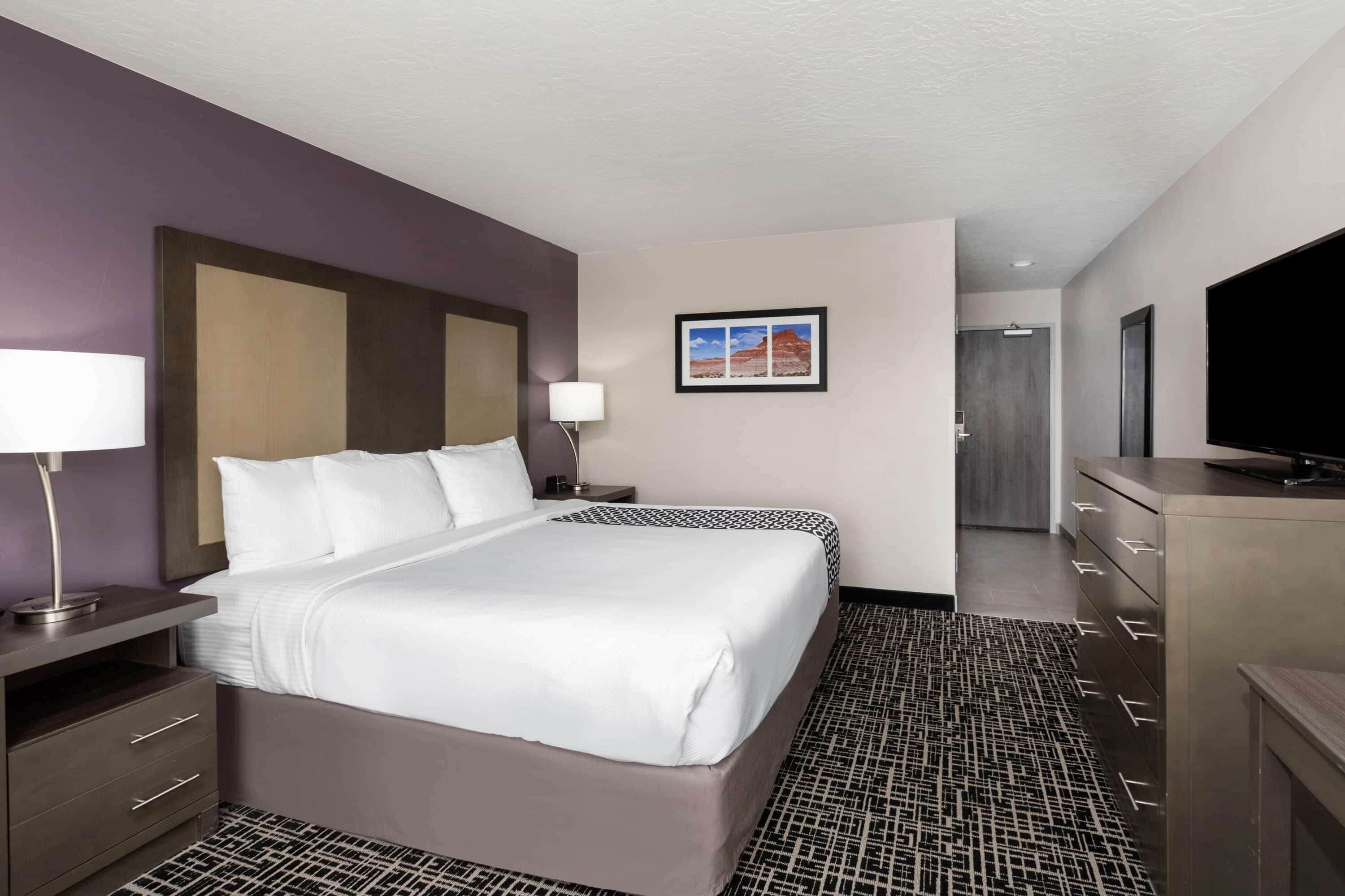 A hotel room with a large bed, a desk with a lamp, a television, and a framed picture on the wall.