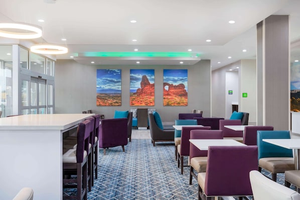 Un moderno vestíbulo de hotel con zona de bar, asientos con sillas moradas y azules, y arte mural con temática desértica.
