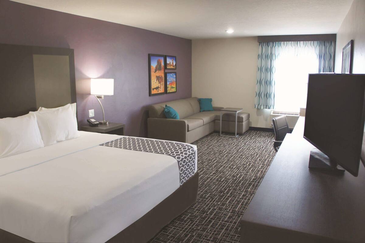 Habitación de hotel con cama, escritorio, silla, televisor, lámpara y ventana con cortinas.