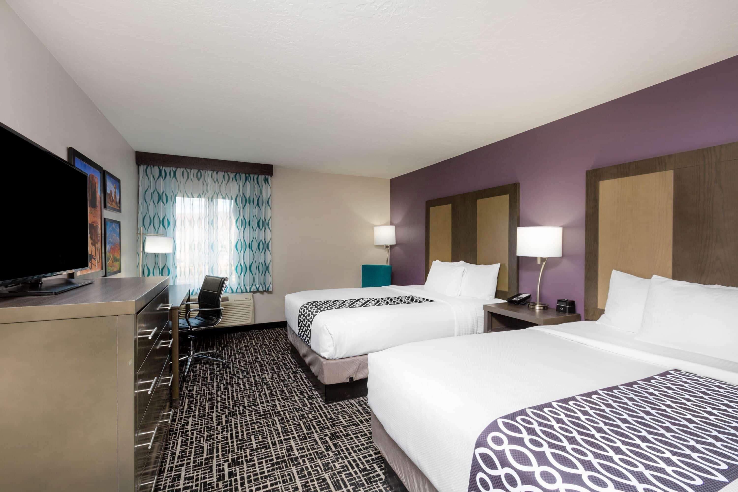 A hotel room with two beds, a desk with a flat-screen TV, a chair, a desk lamp, and a patterned carpet.