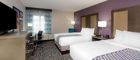 A hotel room with two beds, a desk with a flat-screen TV, a chair, a desk lamp, and a patterned carpet.