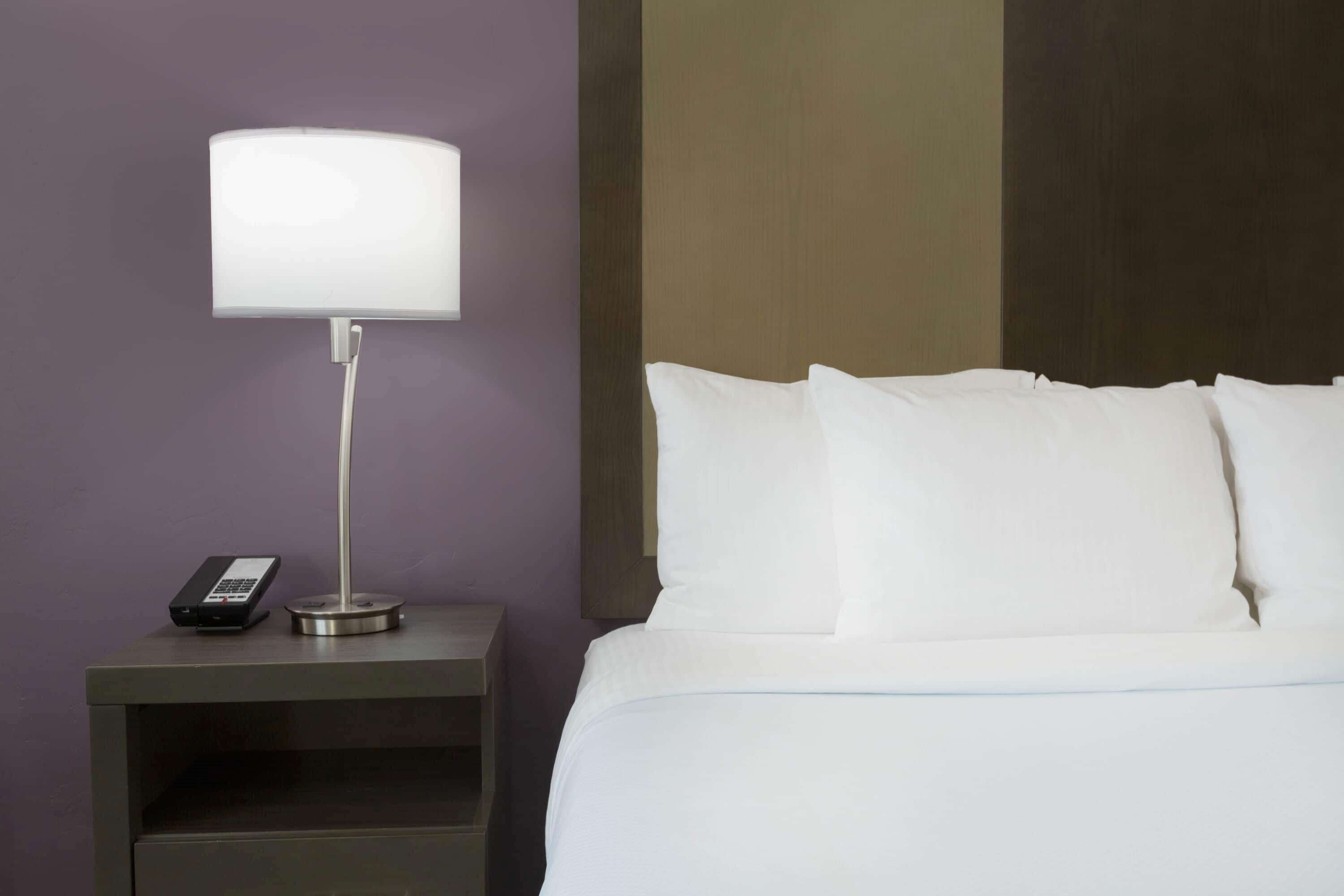 Una cama bien hecha con sábanas y almohadas blancas, una mesita de noche con una lámpara y un teléfono, y una pared con paneles de madera.