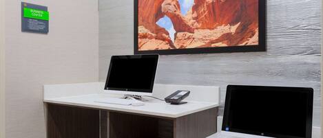 Ein Businesscenter mit zwei Desktop-Computern, einem Drucker und einem an der Wand montierten Fernseher, der eine Schlucht zeigt.