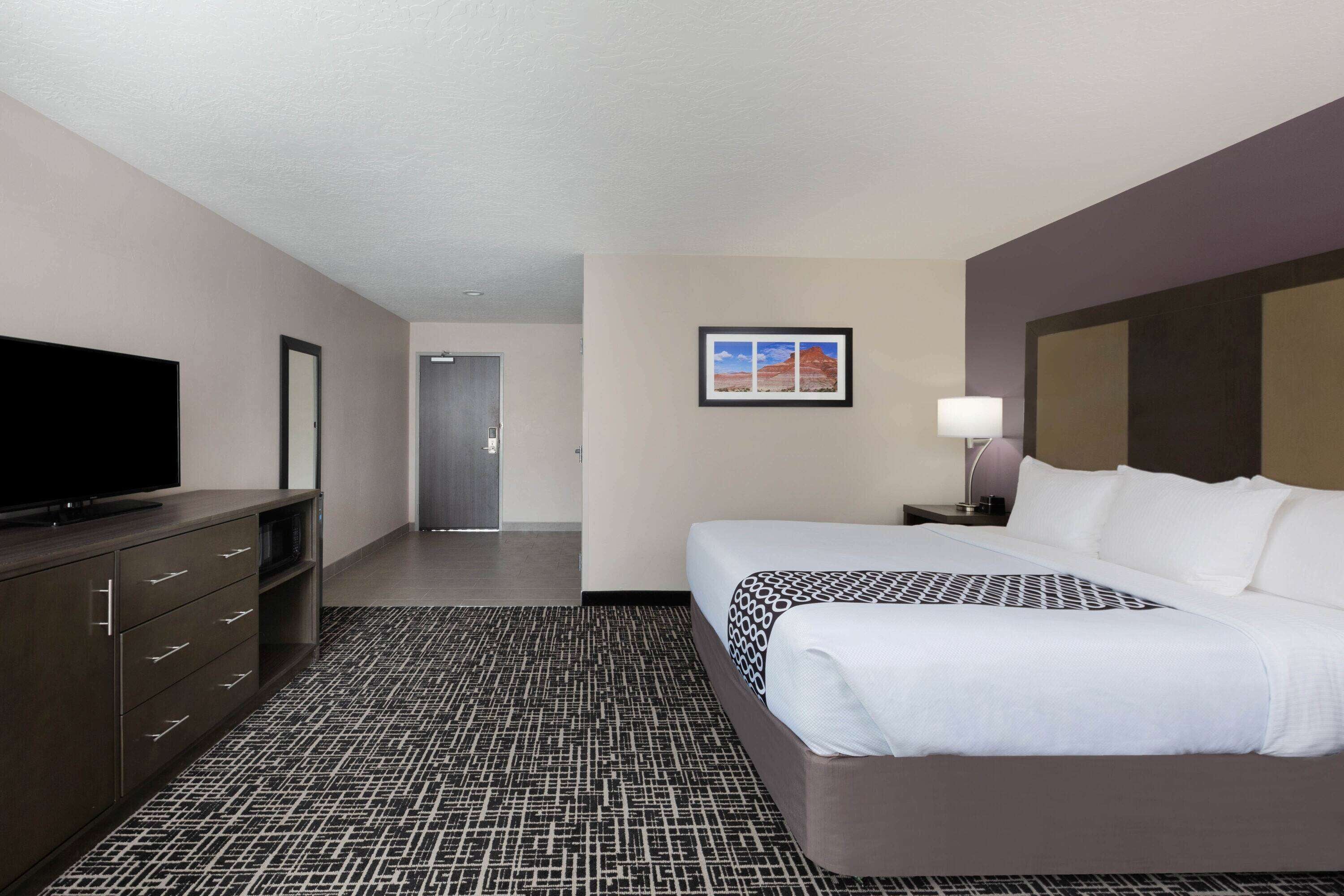 A hotel room with a large flat-screen TV, a bed with white linens, a patterned carpet, and a framed picture on the wall.