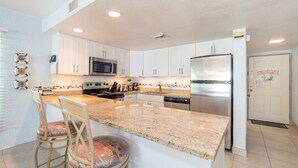 A kitchen with white cabinets, stainless steel appliances, a granite countertop, and two bar stools with cushioned seats.