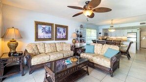 A living room with a sofa, coffee table, and a ceiling fan. There is a kitchen in the background.