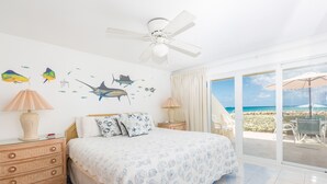 A bedroom with a bed, a nightstand, a lamp, a ceiling fan, and a view of the beach through a sliding glass door.