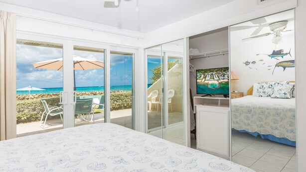 A bedroom with a bed, a TV, and a balcony with a view of the beach and ocean.