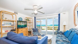 A living room with a TV, a ceiling fan, a sofa, and a view of the beach.