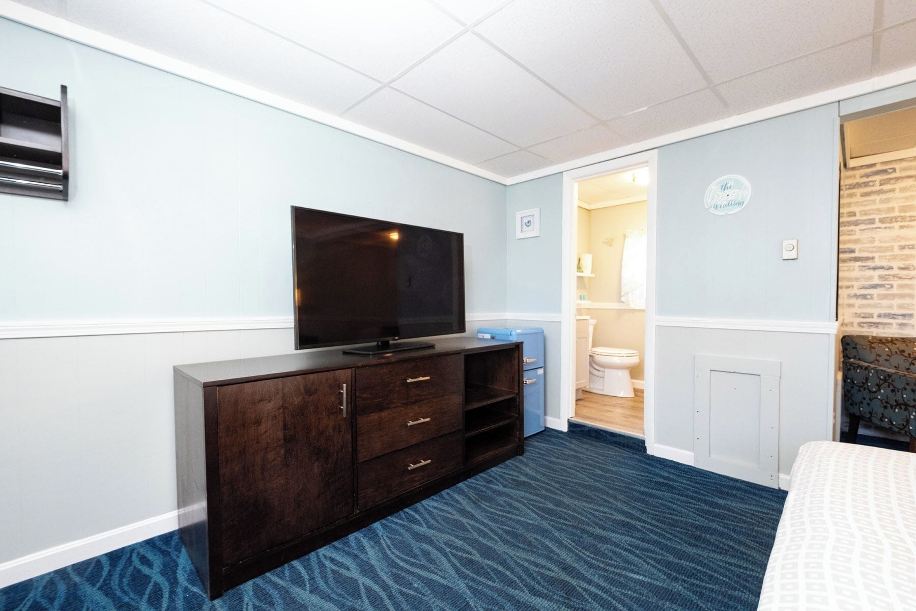 A bedroom with a dark wooden dresser, a flat-screen TV, a blue carpet, and a visible bathroom door.