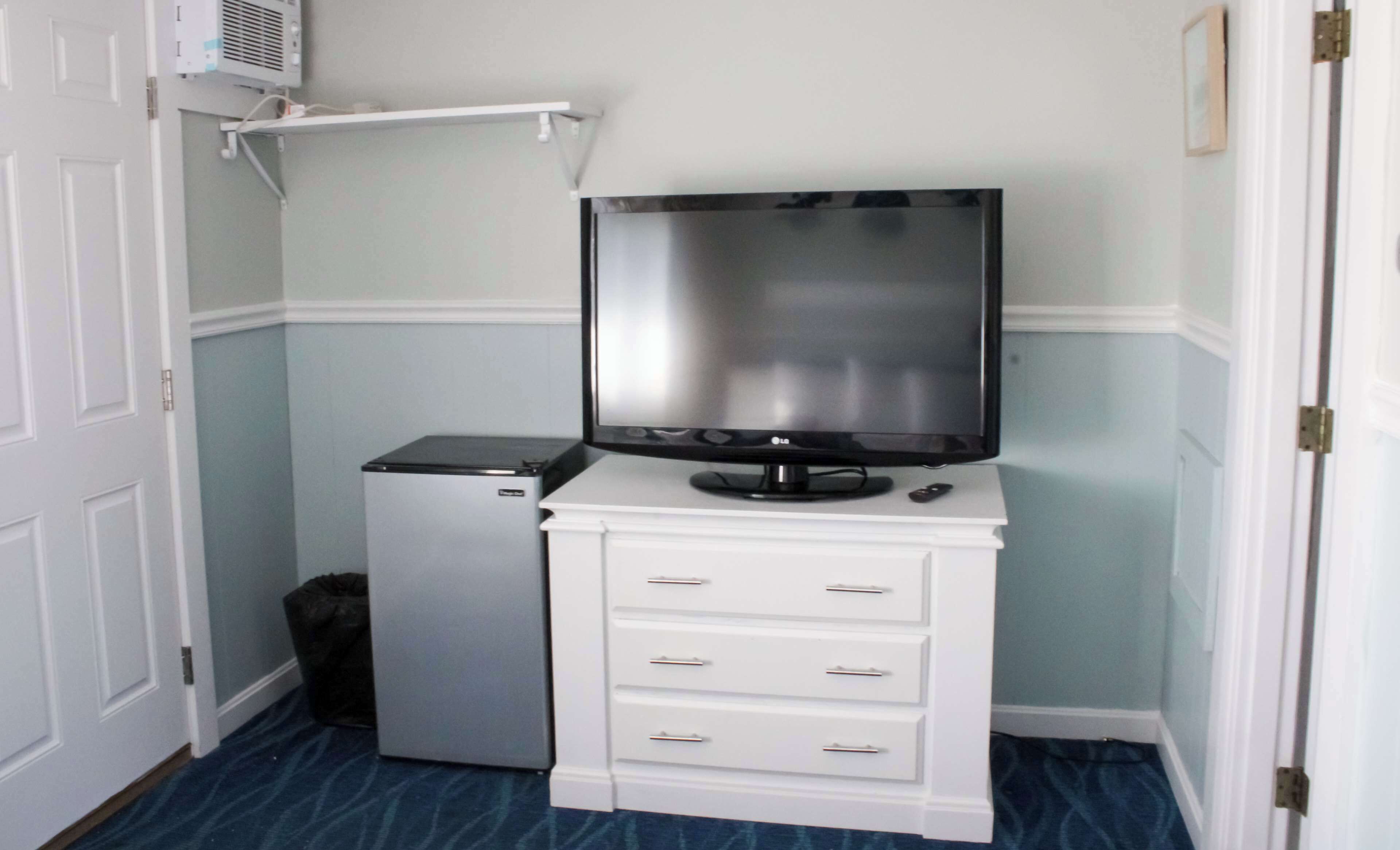 Habitación compacta con un pequeño refrigerador, un televisor de pantalla plana sobre un mueble blanco y un cesto de basura.