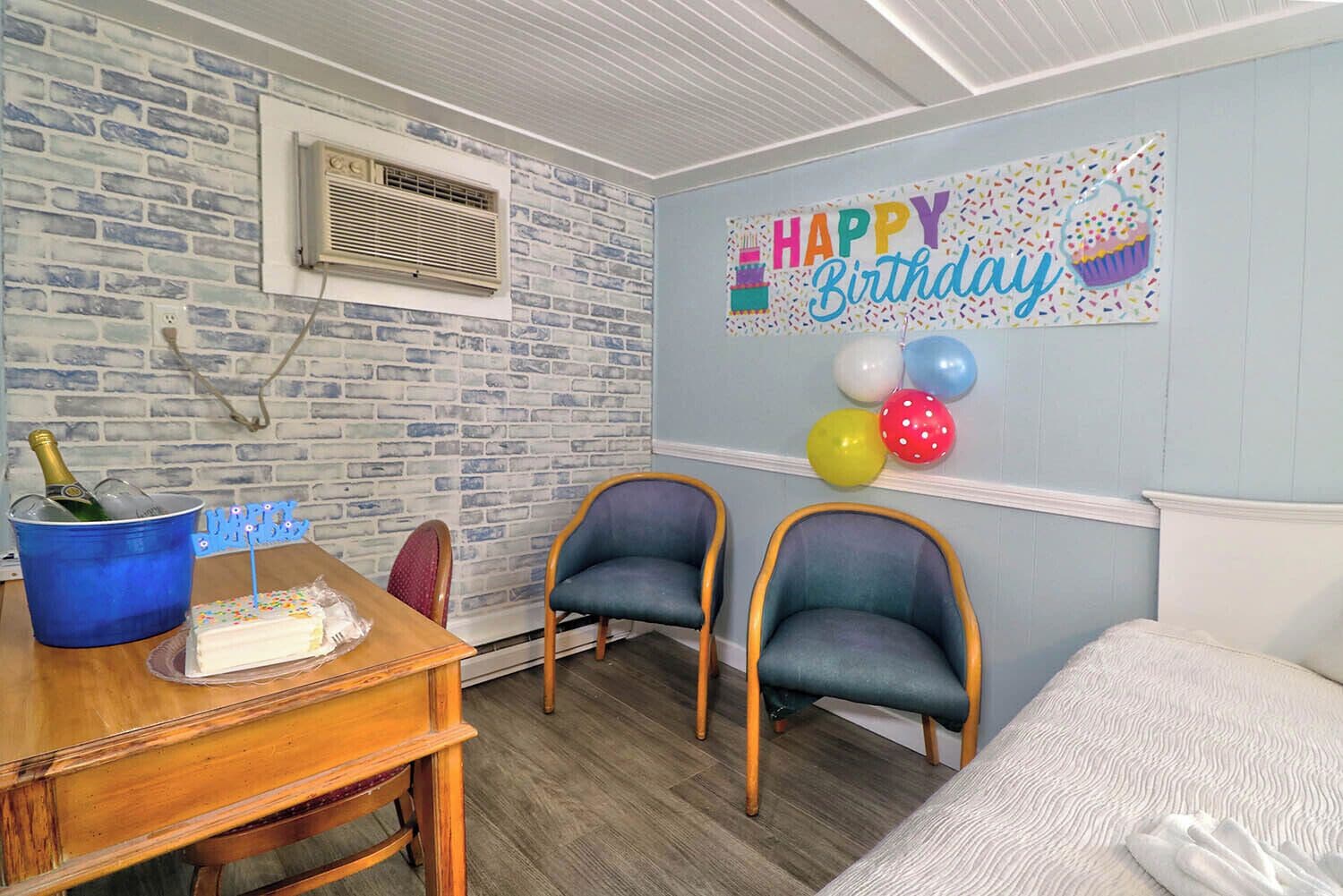 Una habitación con pared de ladrillos, un cartel de 'Happy Birthday', globos, un pastel de cumpleaños y dos sillones.