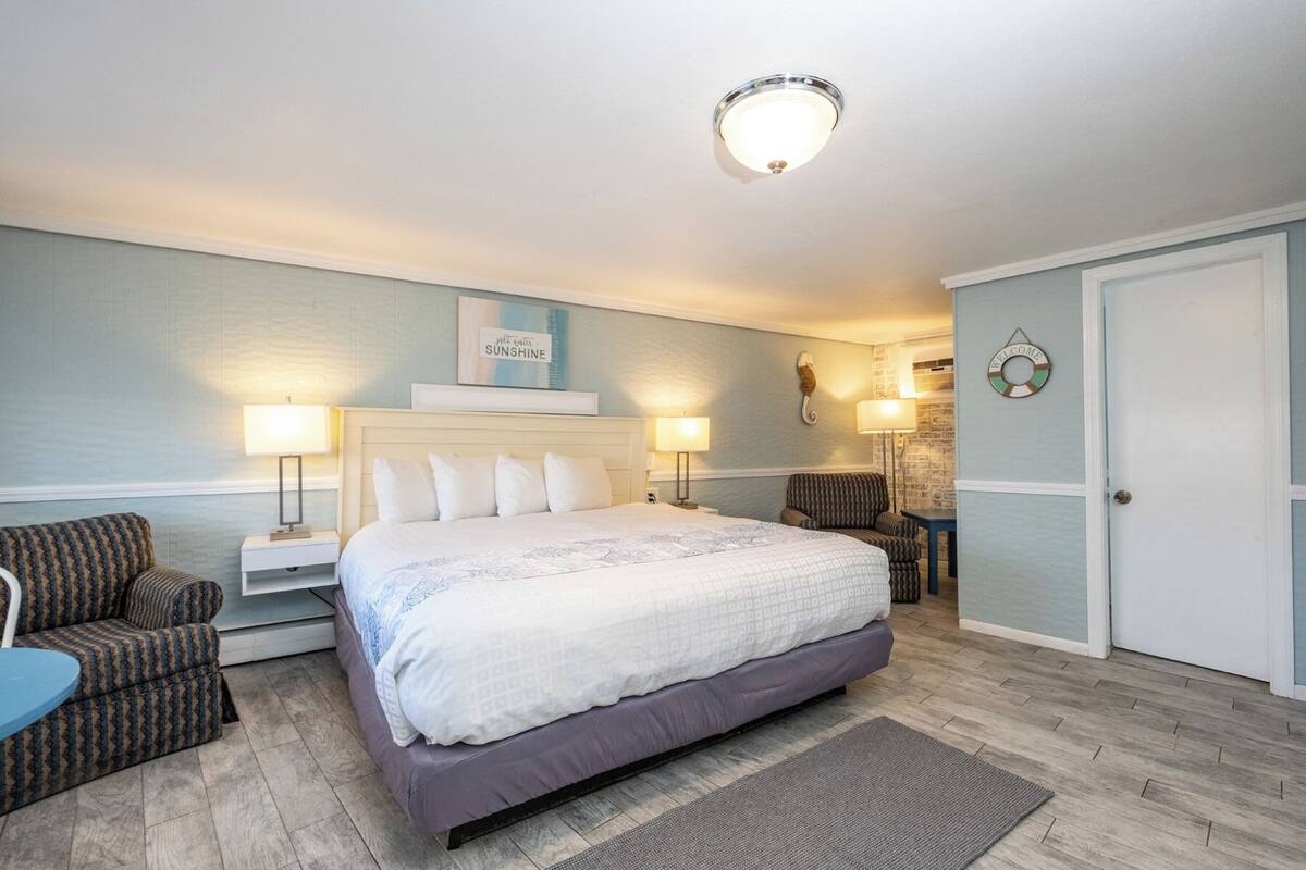 A bedroom with a bed, two bedside tables with lamps, a chair, a rug, and a wall-mounted light fixture.